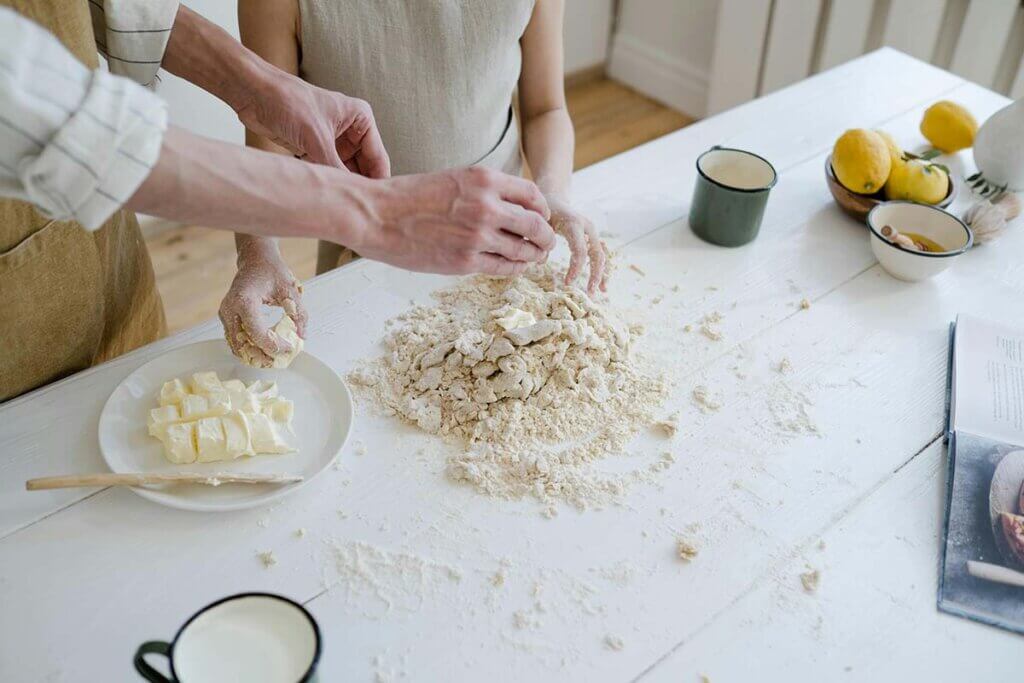 Zwei Menschen backen einen Teig auf einem weissen Holztisch.