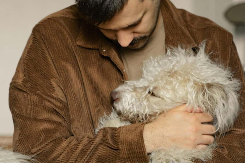 Ein Mann in einer brauen Jacke haelt einen beigen Hund im Arm.