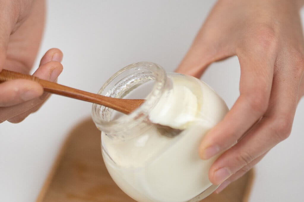 Person haelt ein Glas mit Joghurt in der Hand und holt ein Stueck mit einem Holzloeffel heraus.