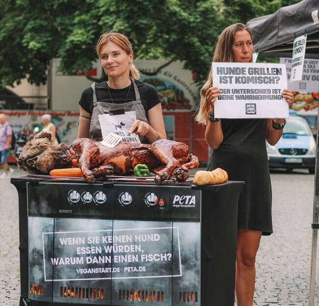 Zwei Personen an einem Grill auf dem eine Hundeattrappe gebraten wird.