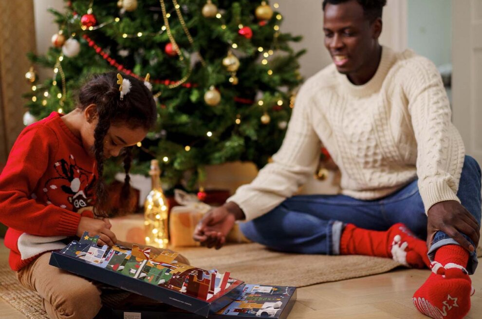 Ein Kind oeffnet einen Adventskalender auf dem Boden vor einem Weihnachtsbaum.