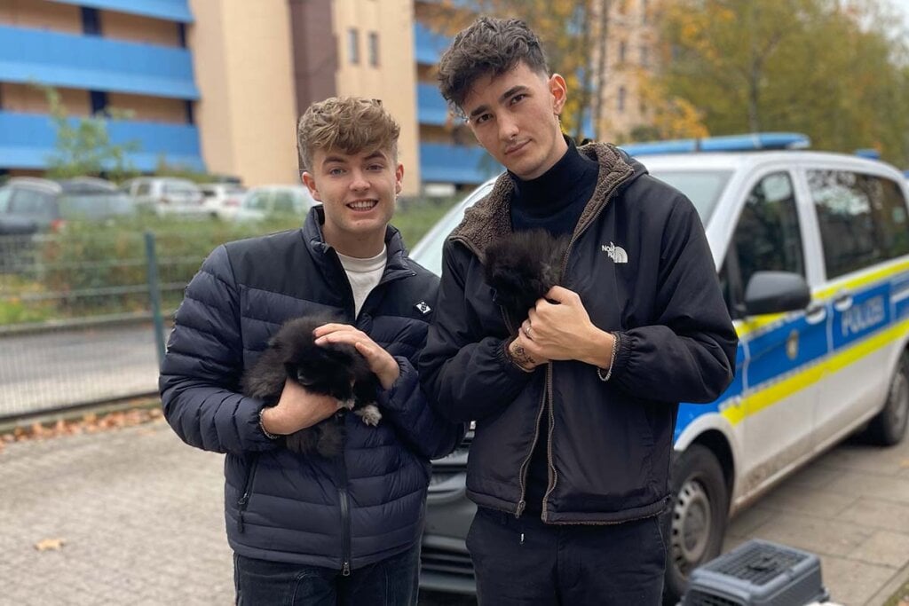 Zwei Maenner stehen mit Welpen in den Armen vor einem Polizeiauto.