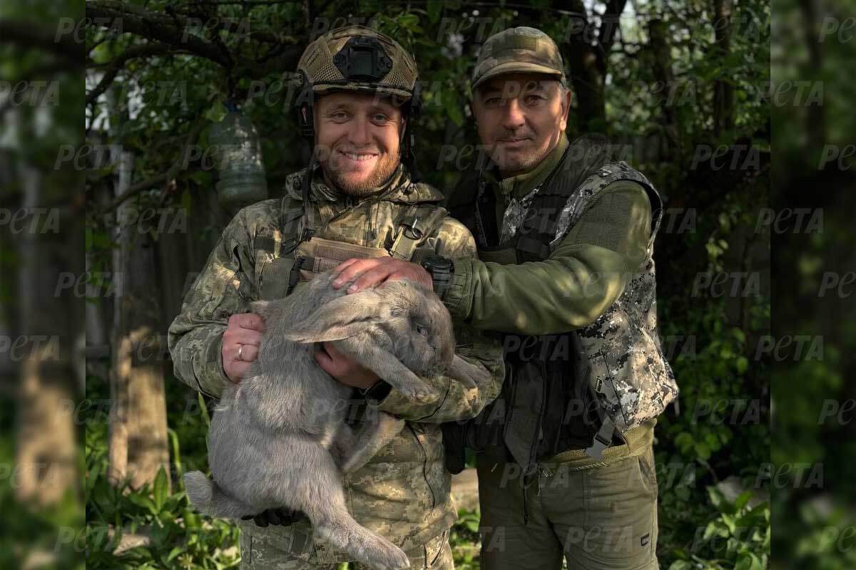 Zwei Männer mit froher Mine schauen in die Kamera, wobei einer von ihnen einen von der Frontlinie geretteten Hasen in den Armen hält.