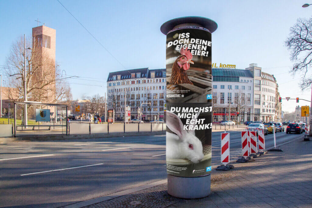 An einer Straße steht im Vordergrund eine Plakatsäule mit Peta-Plakaten.