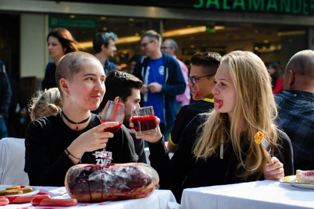 PETA Streetteam Demonstration gegen Fleisch. Zwei Personen sitzen an einem Tisch und stossen mit Glaesern an, in denen eine rote Fluessigkeit ist.