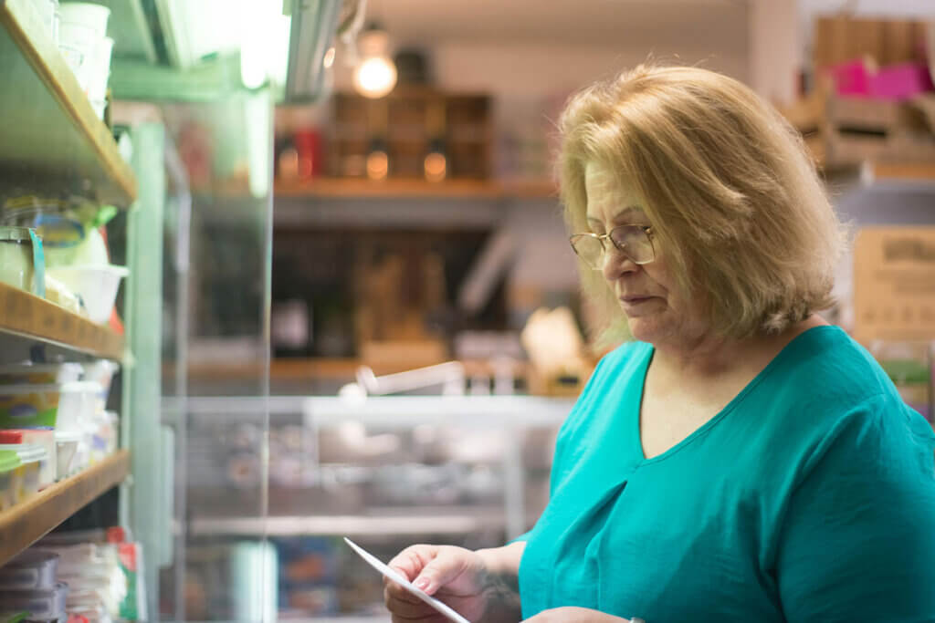 Eine Frau im gruenen Hemd steht vor einem Supermarktregal und liesst einen Zettel.